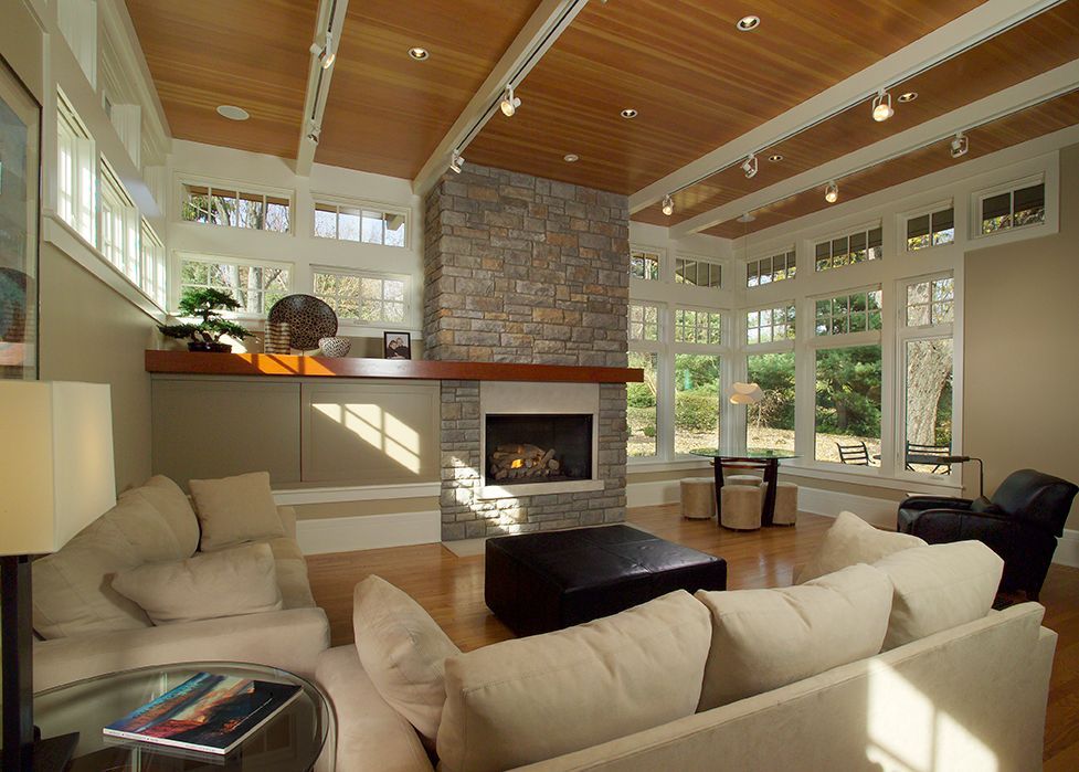 Spacious living room with stone fireplace, large windows, light-colored couches, wood ceiling, and a large black ottoman.