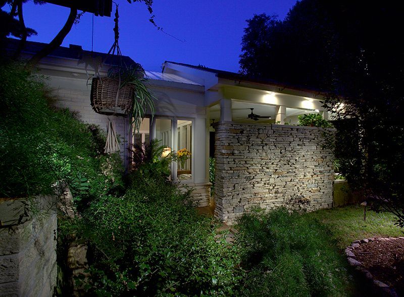 Night view of a white house with stone wall illuminated by warm light. Lush greenery surrounds the house.