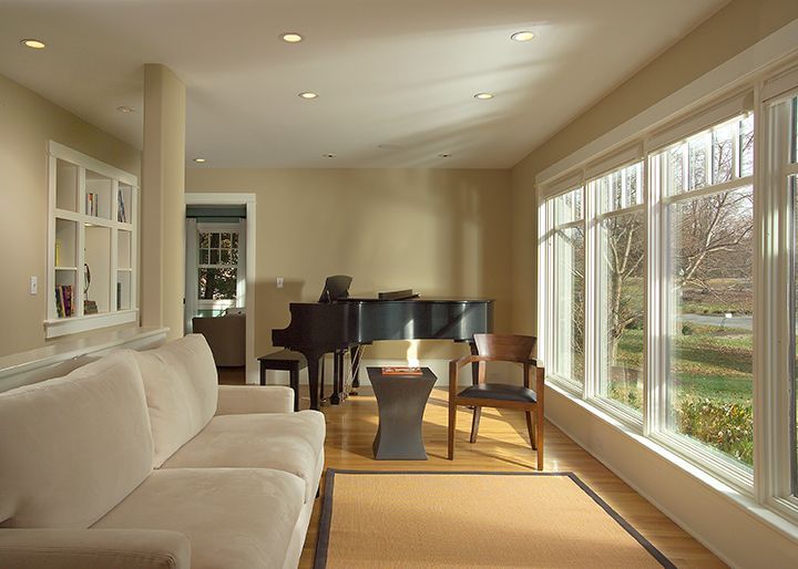 Living room with piano, sofa, and large windows; light beige walls and hardwood floors.