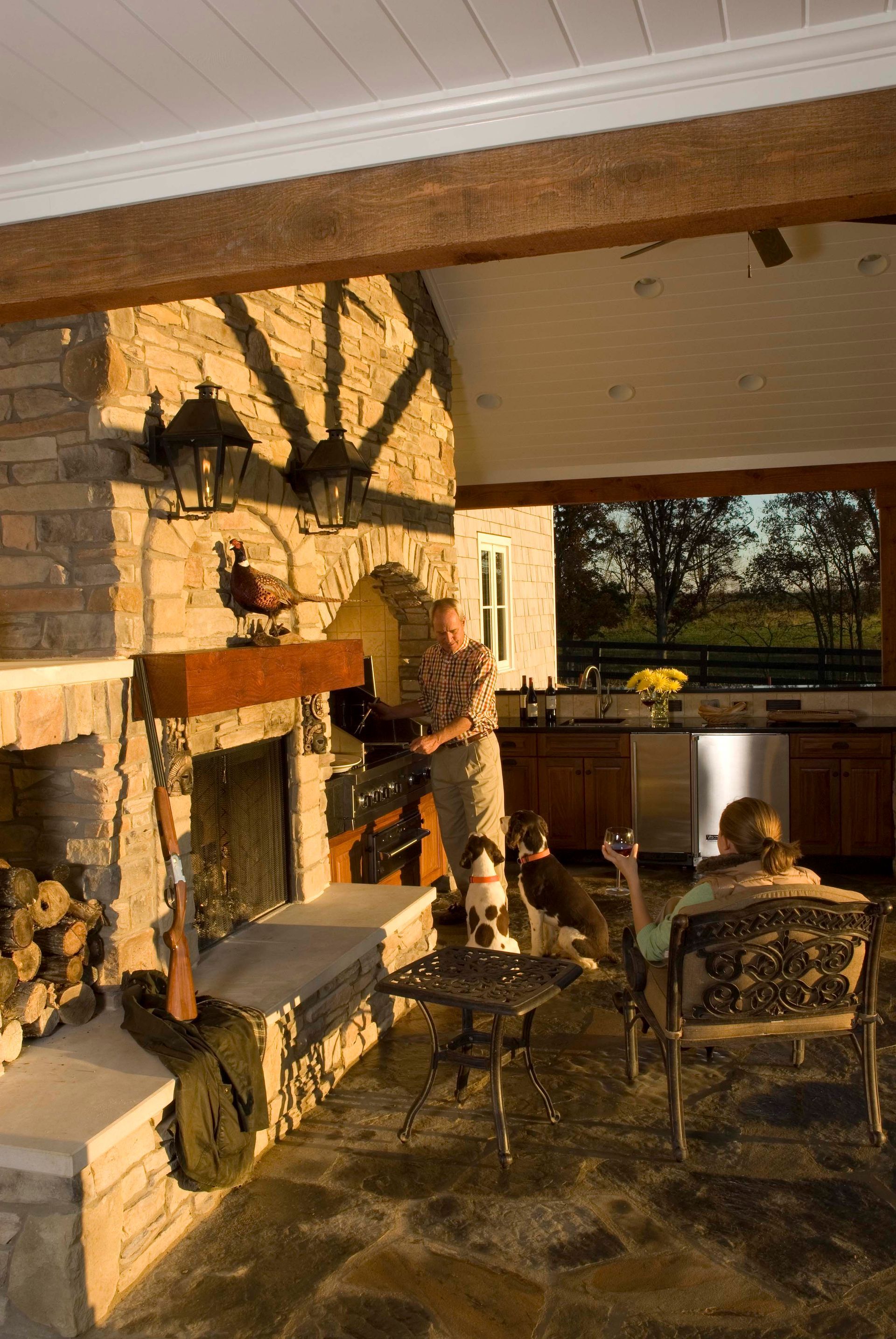 Man placing wood in stone fireplace, woman relaxing, dog. Outdoor kitchen with stone and wood beams.