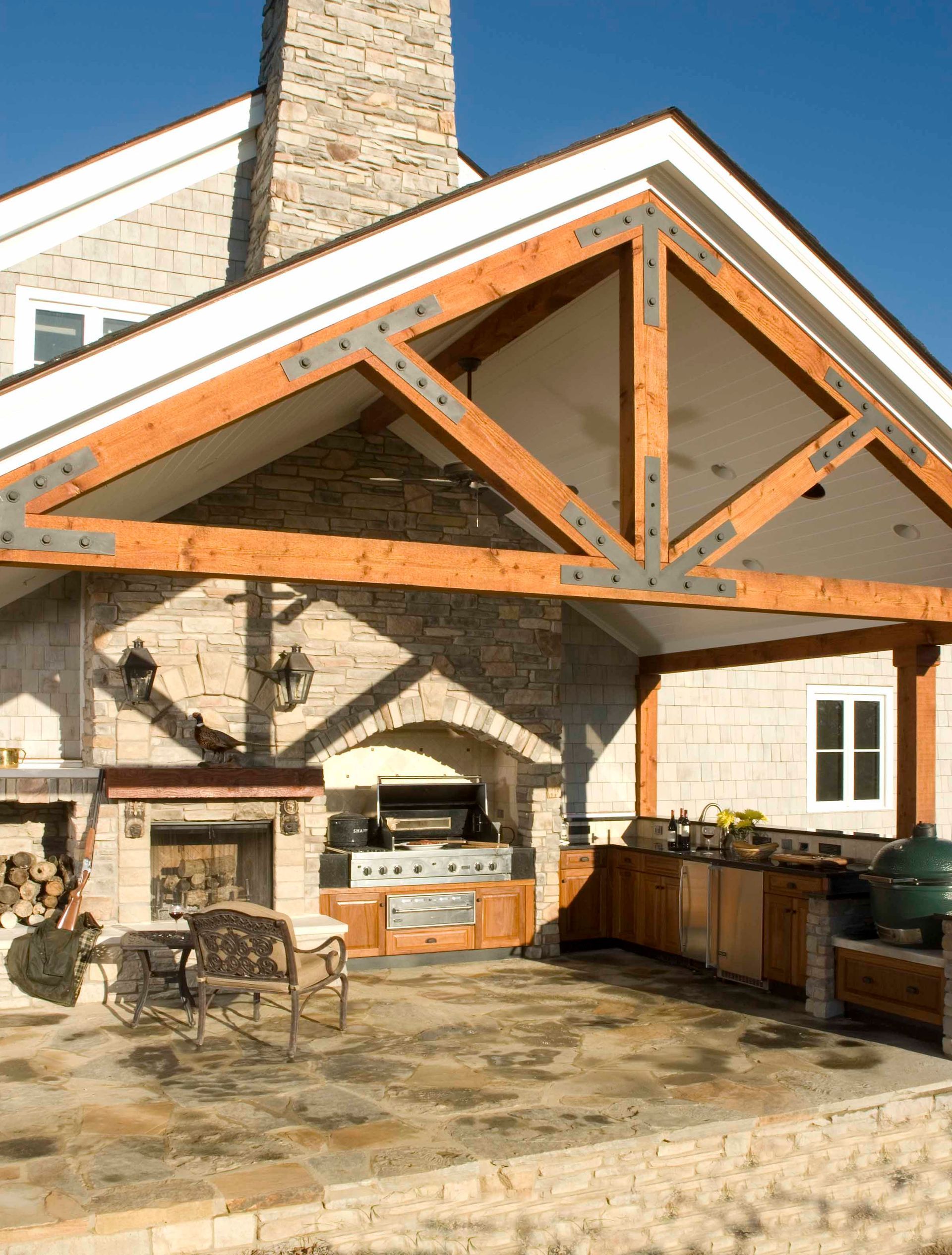Outdoor kitchen with fireplace, grill, wooden beams, stone walls, and cabinetry.