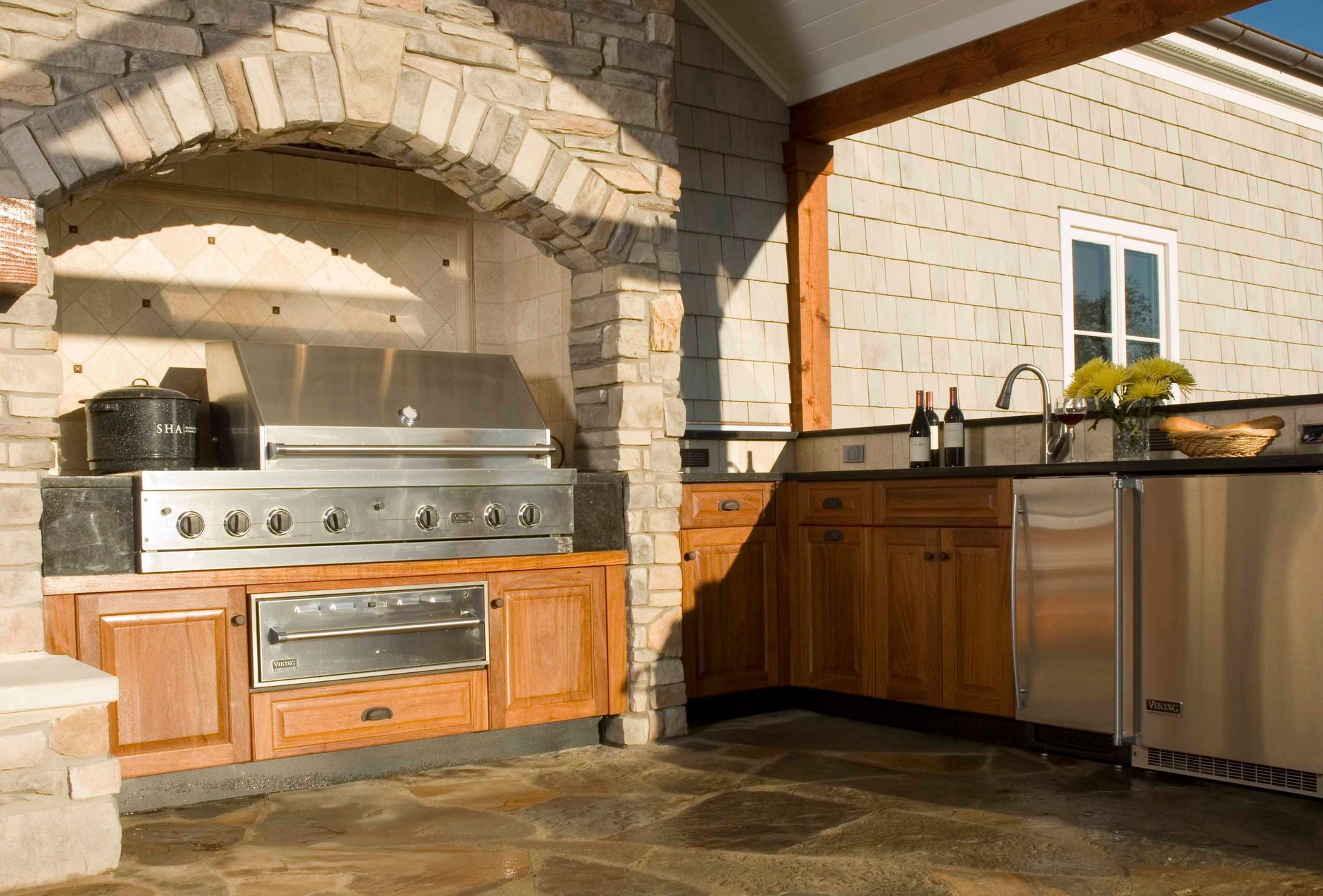 Outdoor kitchen with stone grill, stainless steel appliances, and wooden cabinets.