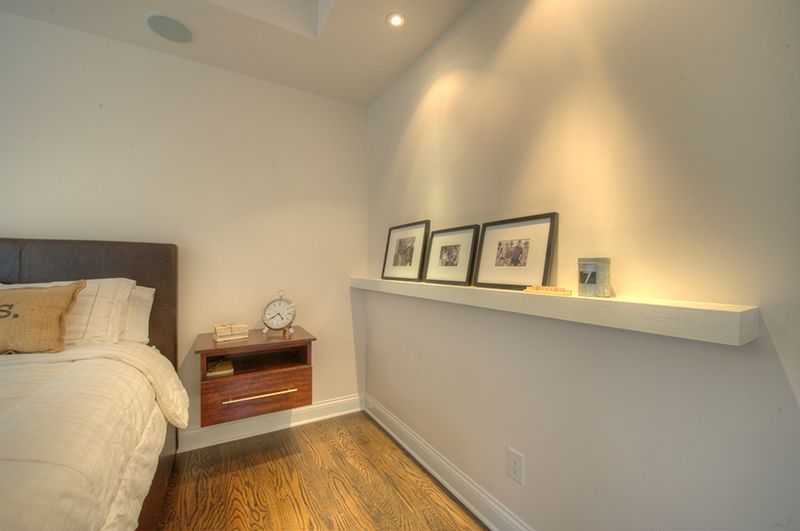 Bedroom with floating shelf, pictures, nightstand, and a bed. White walls, dark wood floors.