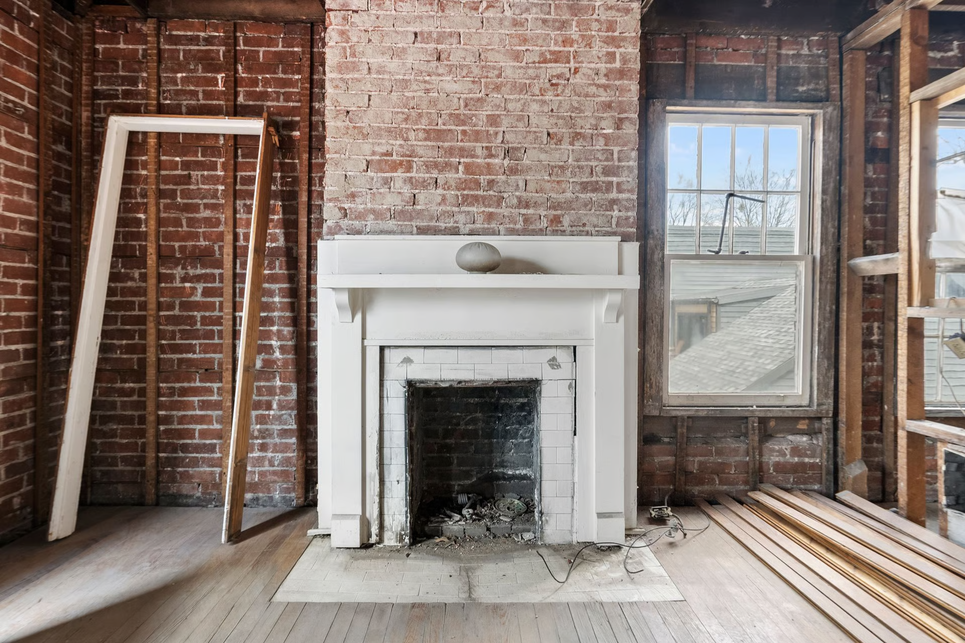 Fireplace in a room under renovation, exposed brick, window, and framing.