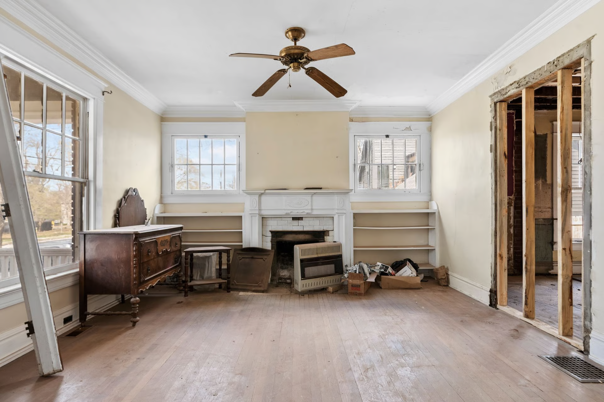 Empty room with a damaged fireplace, windows, and wood floors, suggesting renovation.