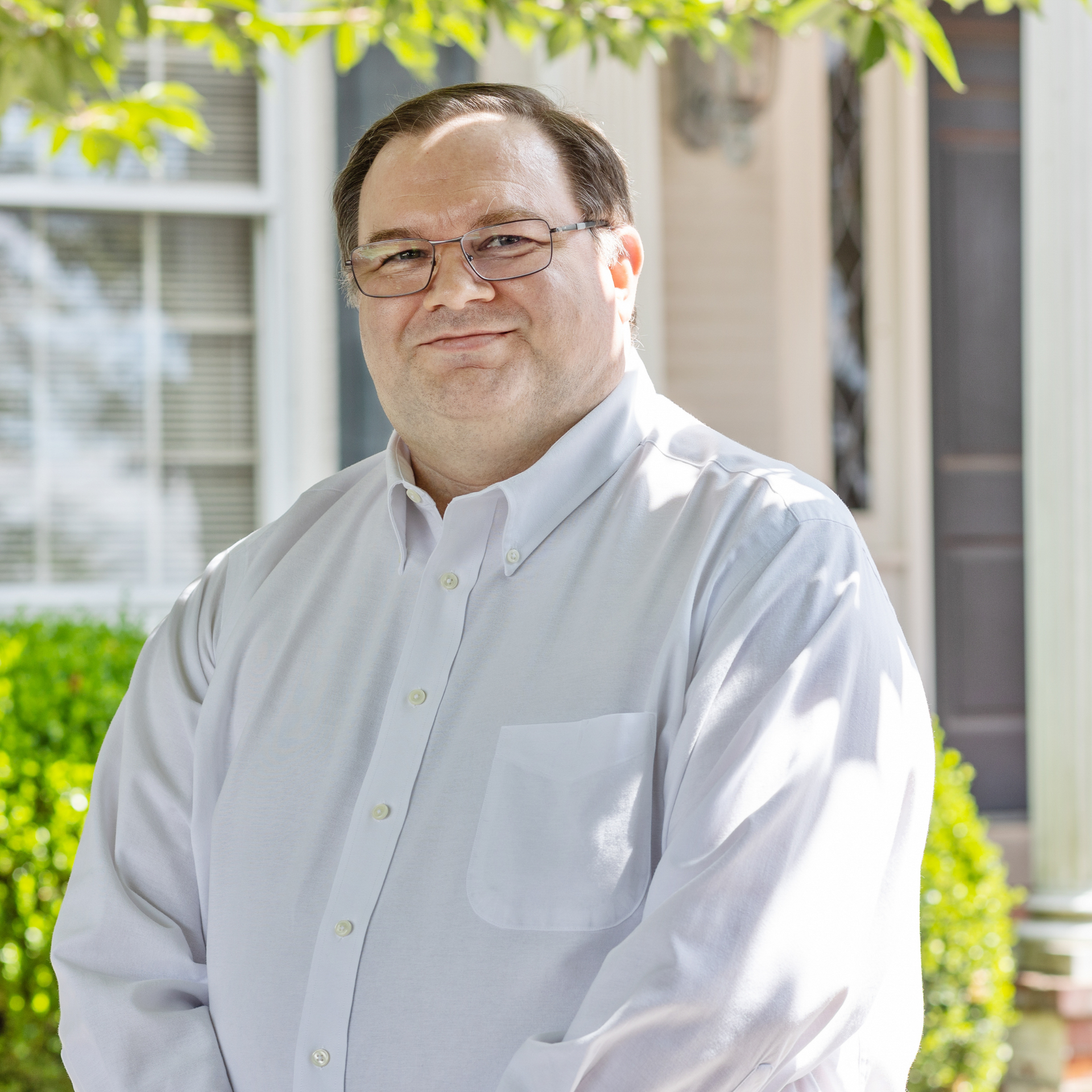 Man in glasses and white shirt smiles outside a building with greenery in the background.