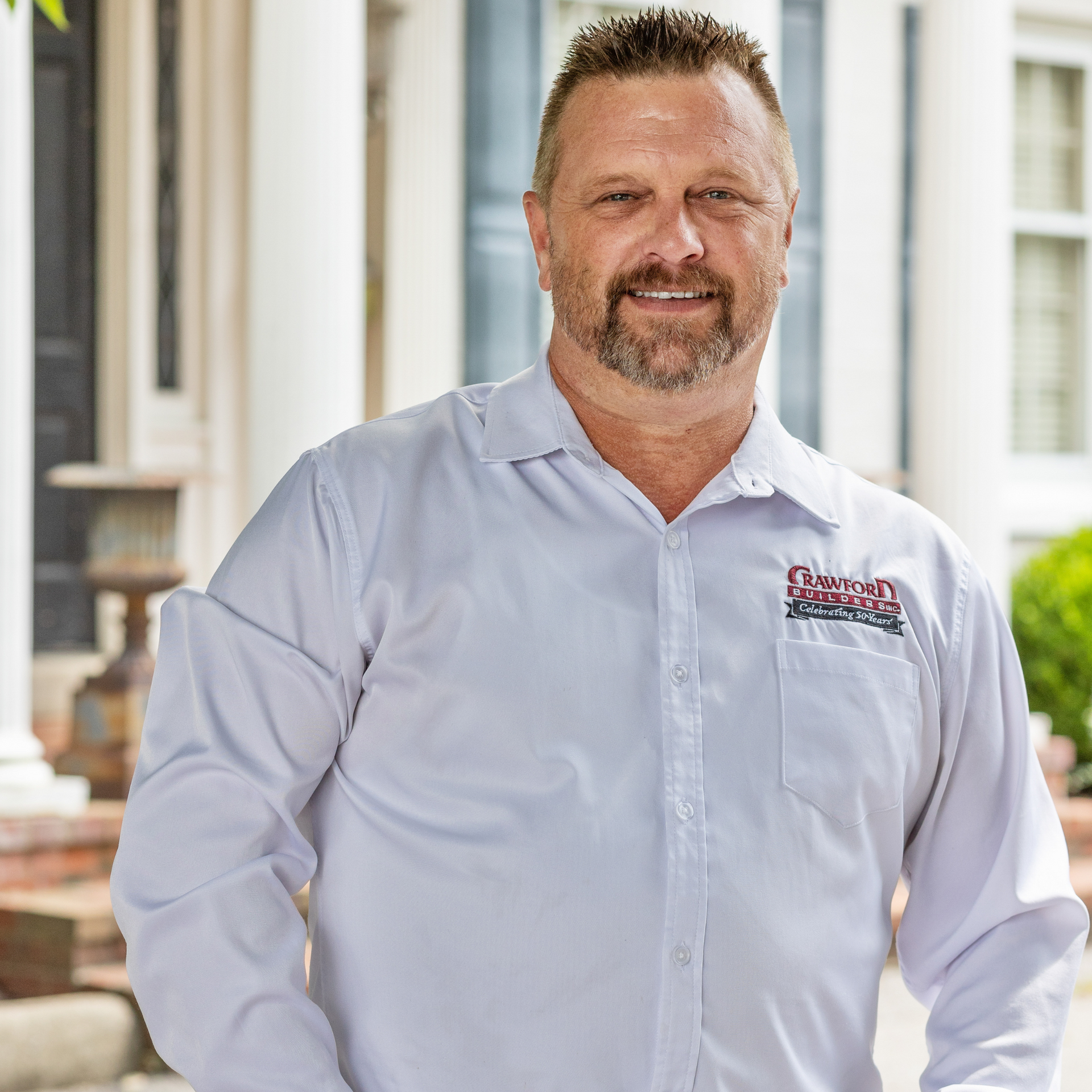 Man with a beard, wearing a white shirt with a logo, smiles in front of a building.