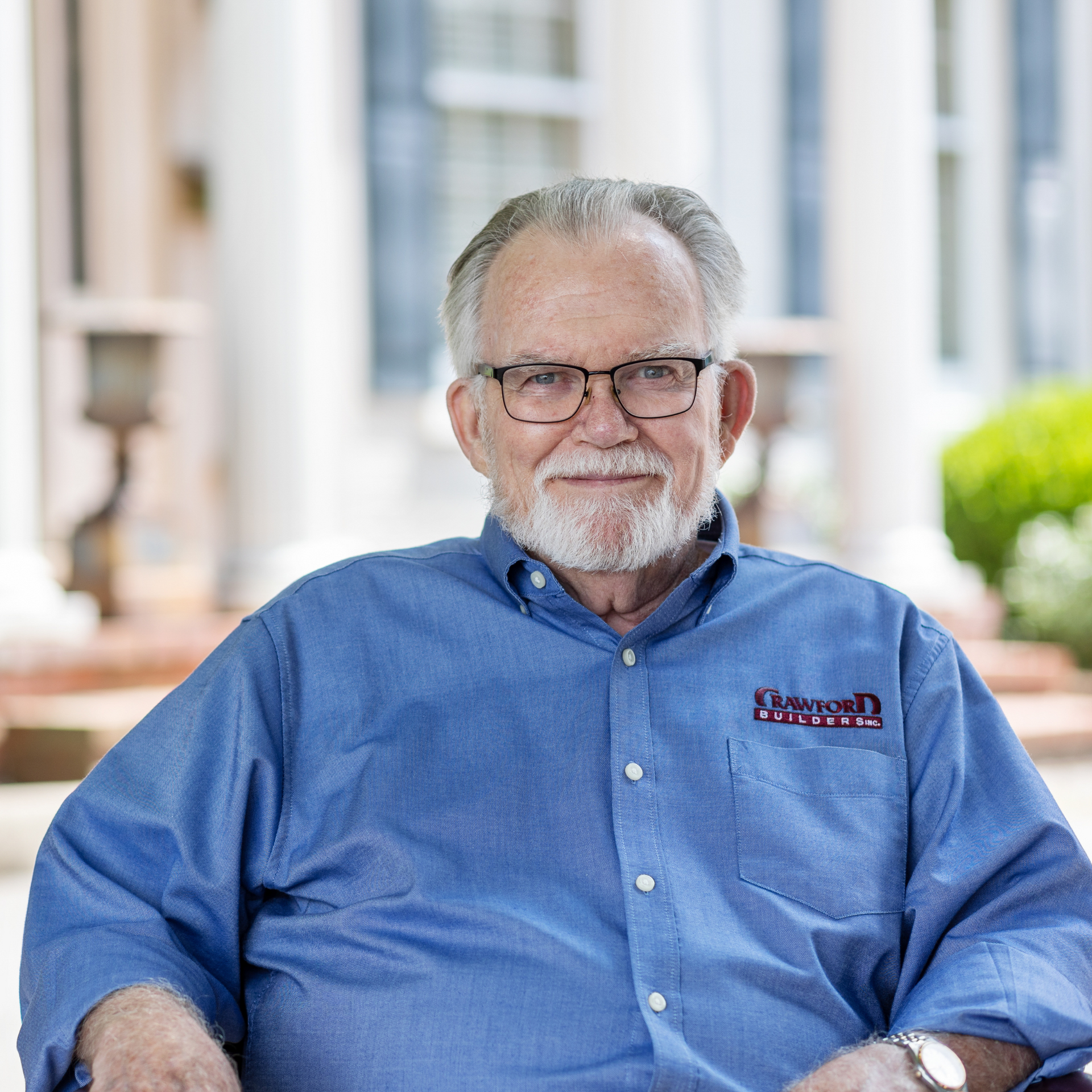 Elderly man with glasses, blue shirt, and gray beard smiles outdoors.