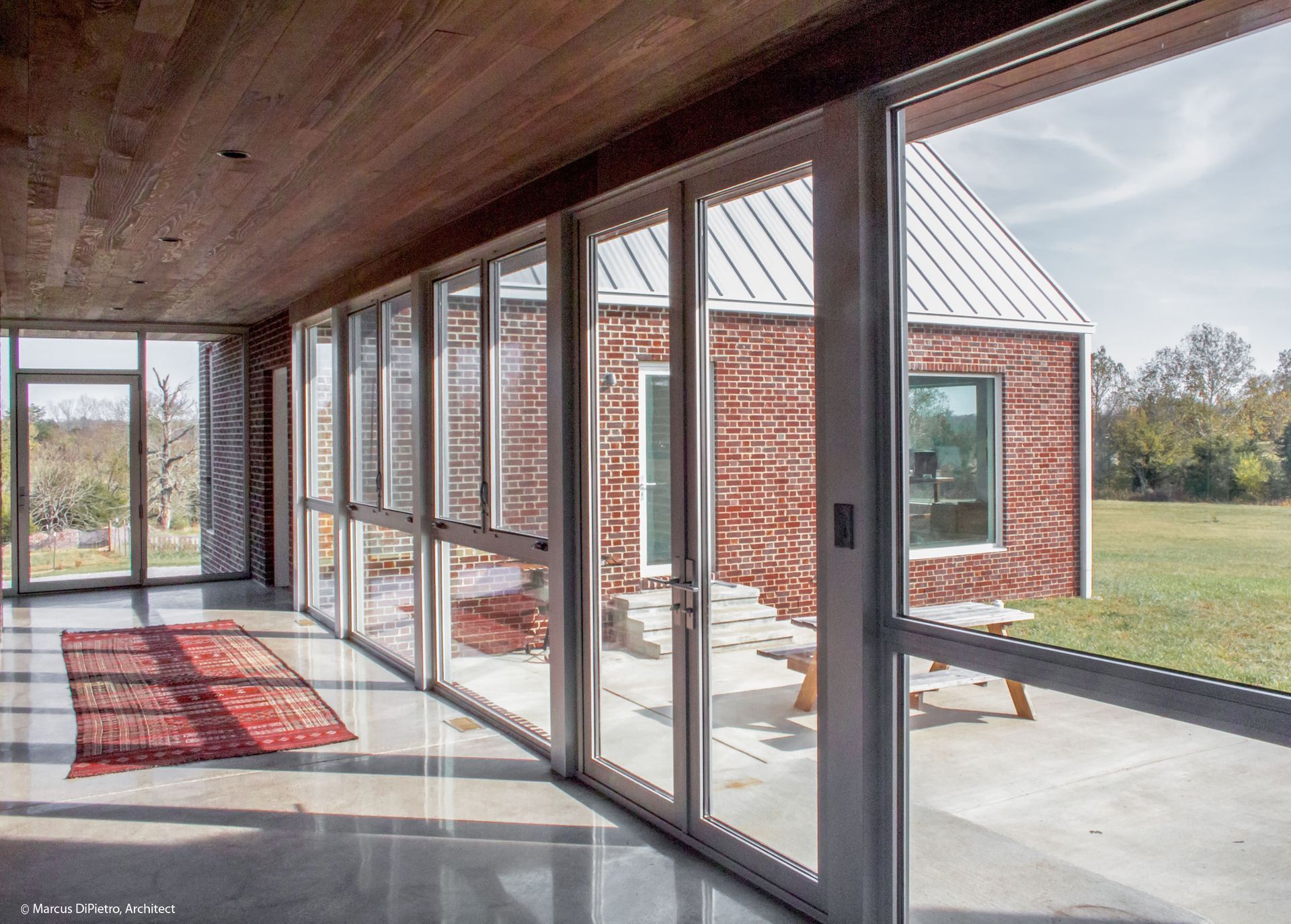 Interior view with glass doors, red rug, and brick building with metal roof on the outside, overlooking a field.