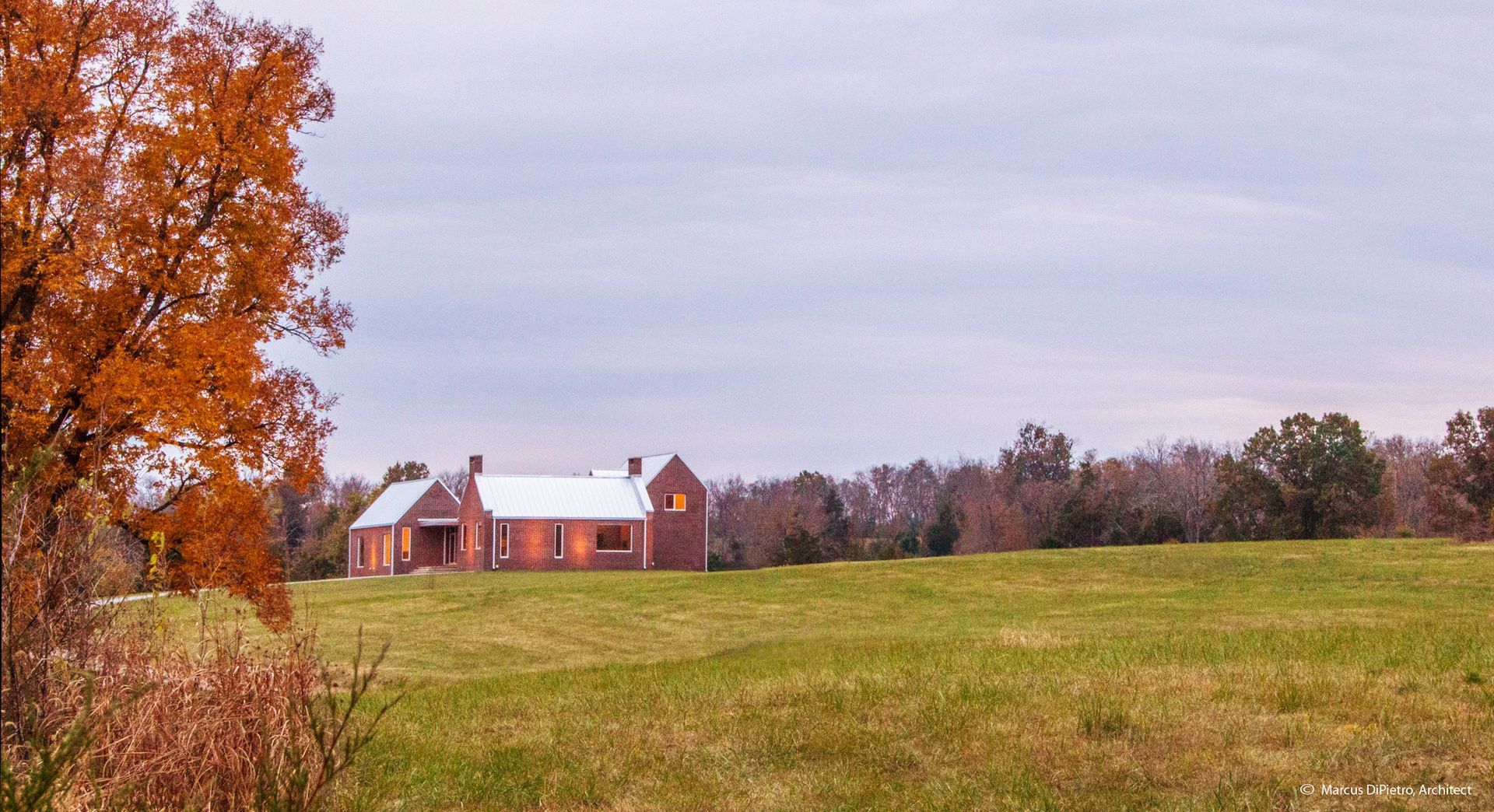 Autumnal scene: Brick house in a field with a tree displaying orange leaves under a cloudy sky.