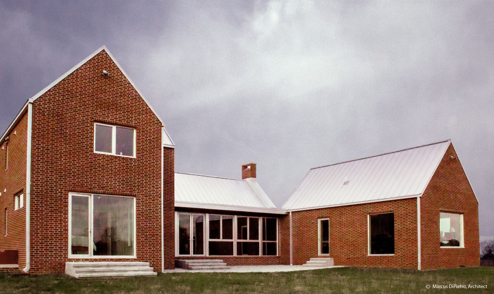 Brick house with gabled roofs, large windows, and a metal roof, set on green grass under a cloudy sky.