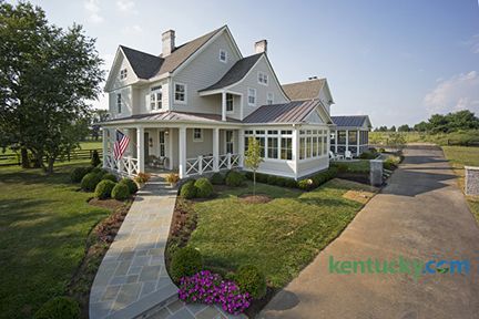 Light gray farmhouse with porch, American flag, landscaped yard, and long driveway.