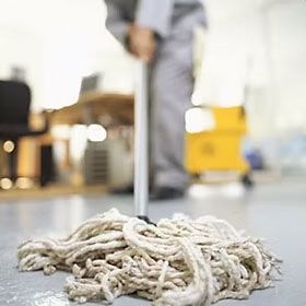 Mop head on floor, blurred person mopping in the background. White mop, gray floor, office setting.