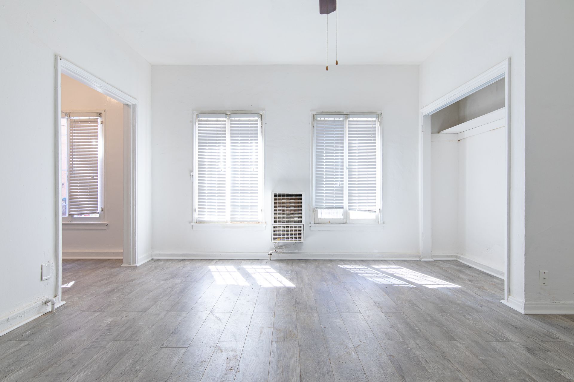 Empty, bright living room with two windows, wood floors, and doorways.