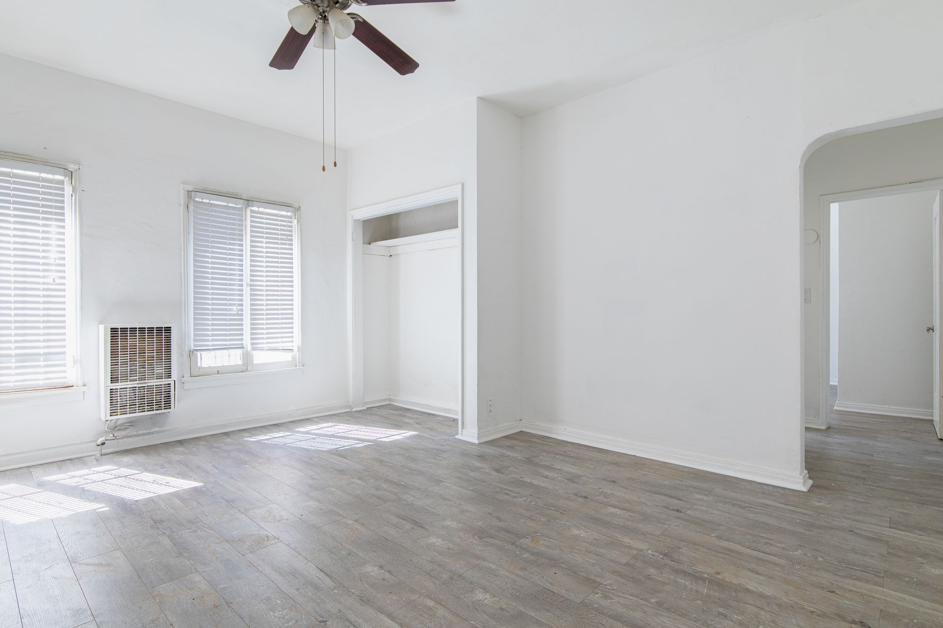 Empty apartment room with gray wood floors, white walls, two windows, and closet.