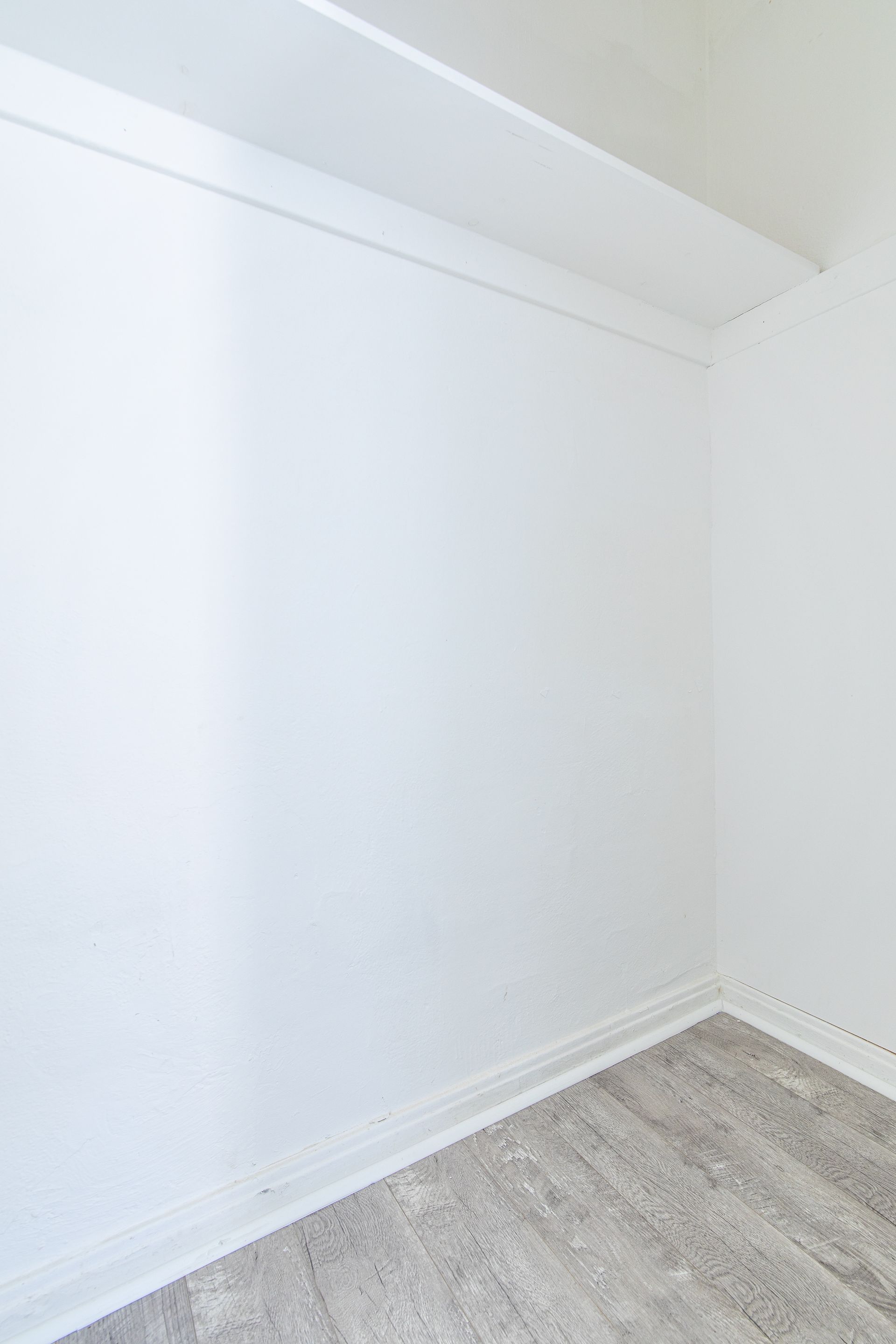 Empty white-walled closet corner with a shelf at the top and gray wood-look flooring.