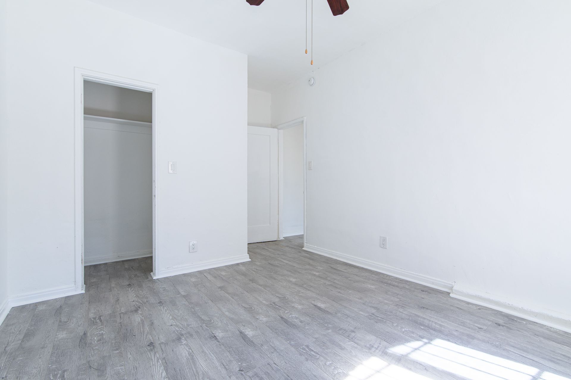 Empty bedroom with grey wood-look flooring, white walls, closet, and a ceiling fan.