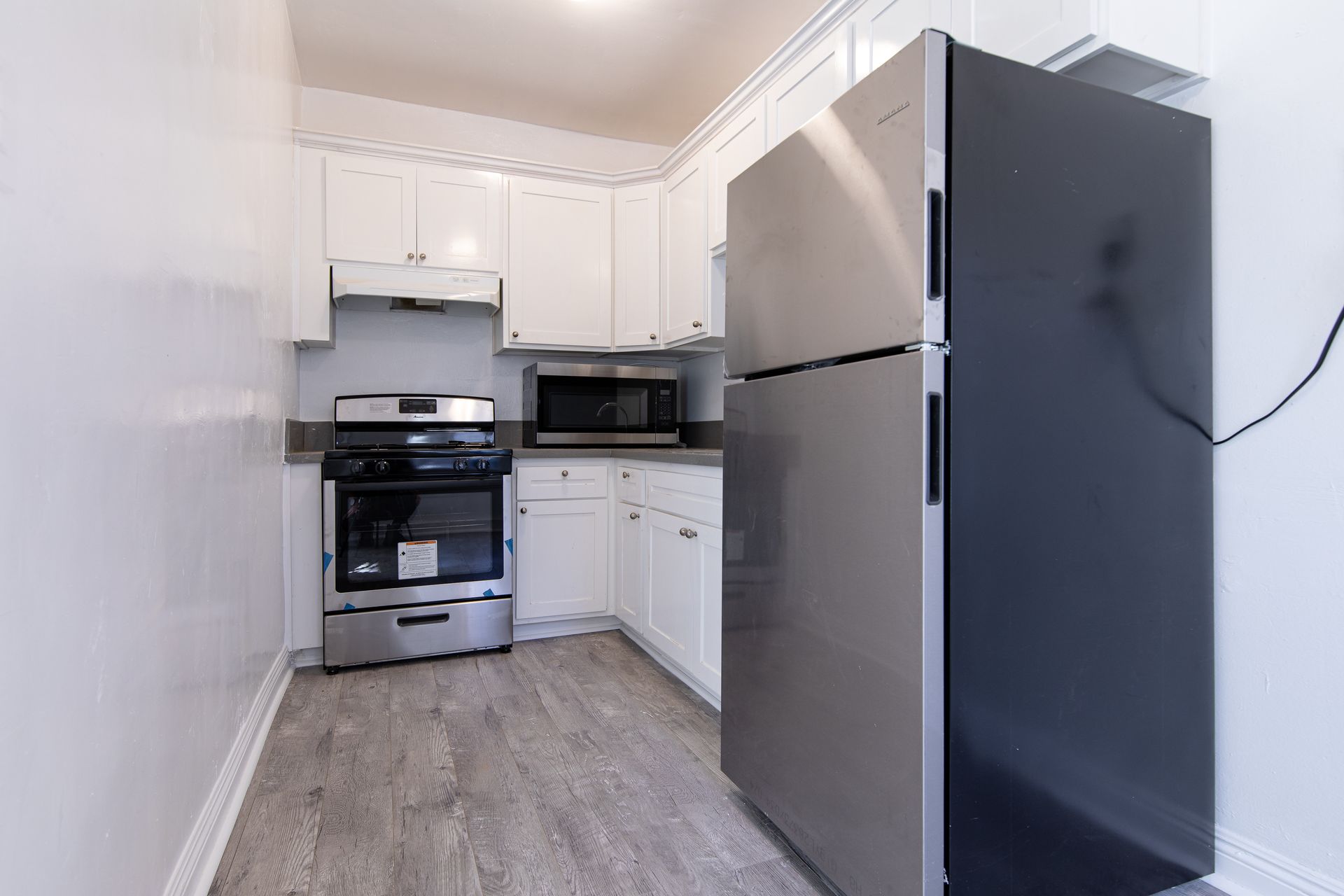 Small, white kitchen with stainless steel appliances: fridge, stove, microwave, and cabinets. Gray flooring.