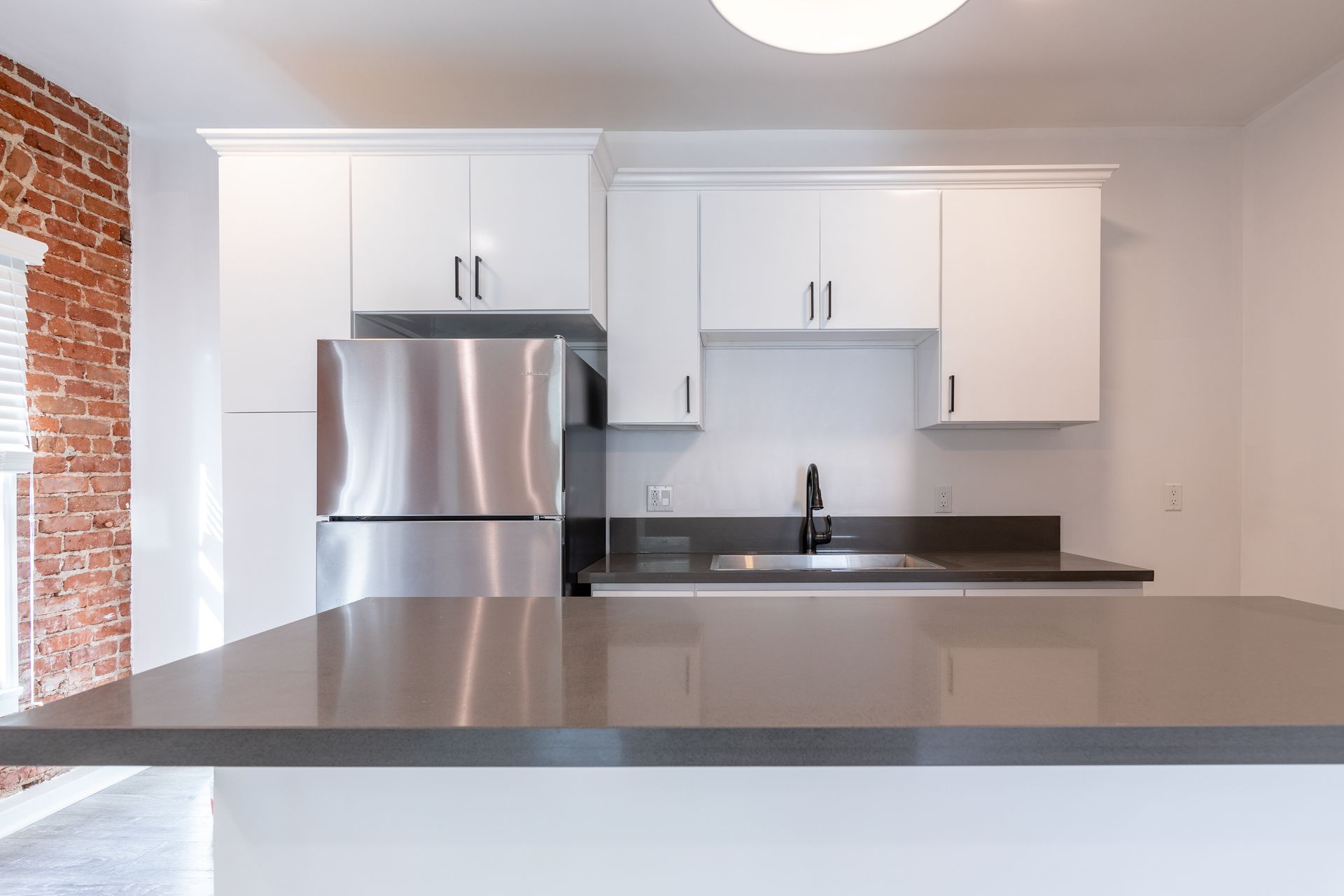 Modern kitchen with white cabinets, stainless steel refrigerator, and gray countertop. Exposed brick wall on the left.