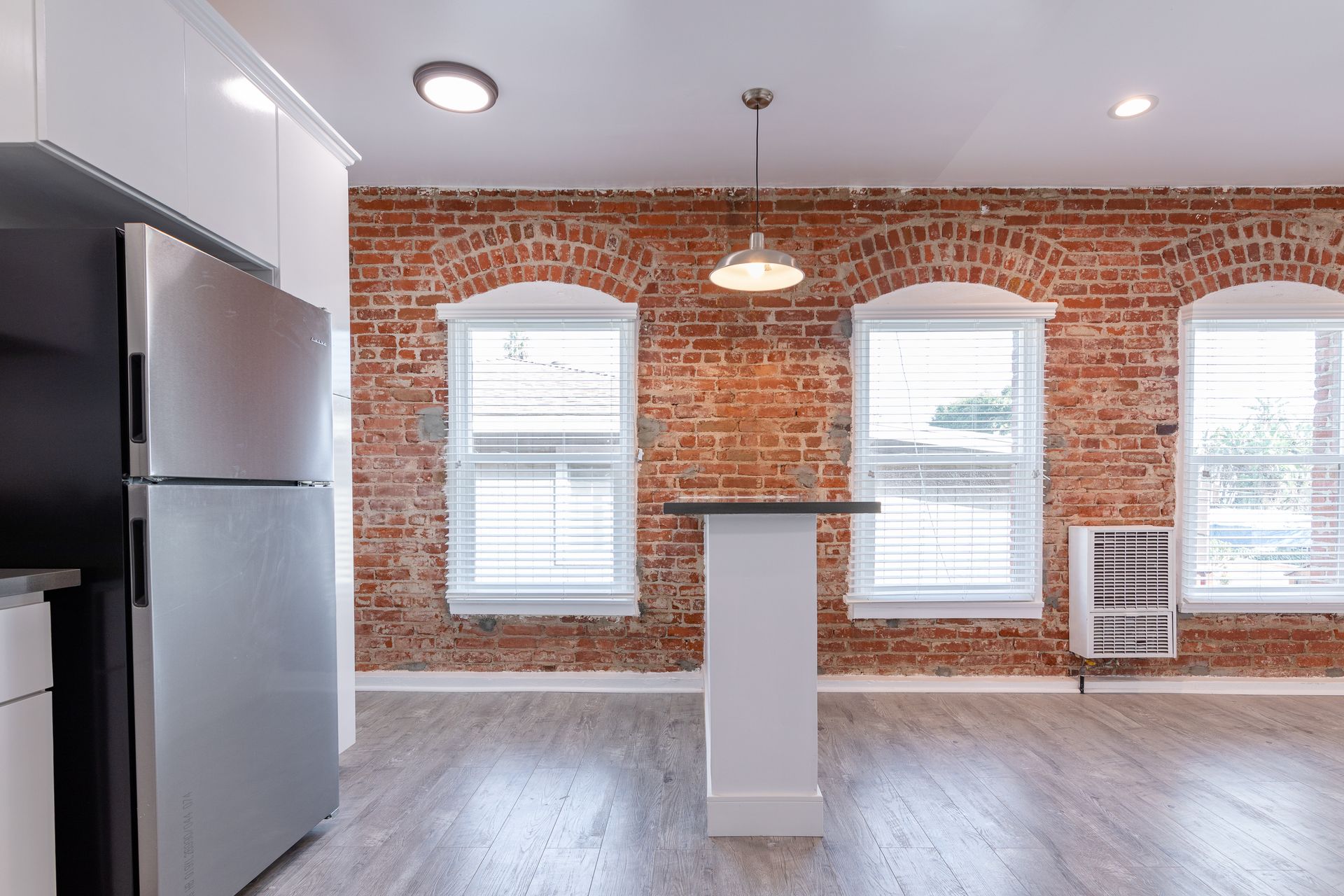 Brick wall interior with windows, stainless steel refrigerator, white cabinets, and a kitchen island.