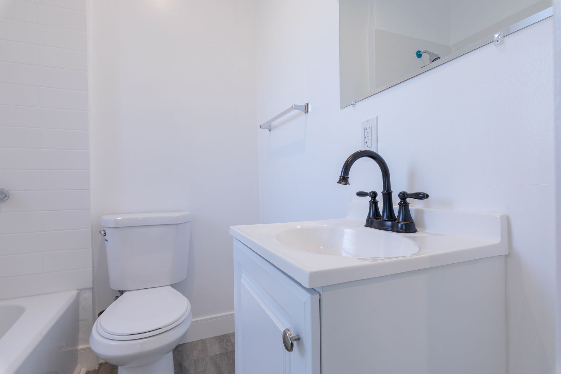 White bathroom with toilet, sink, and mirror. Black faucet and towel rack.