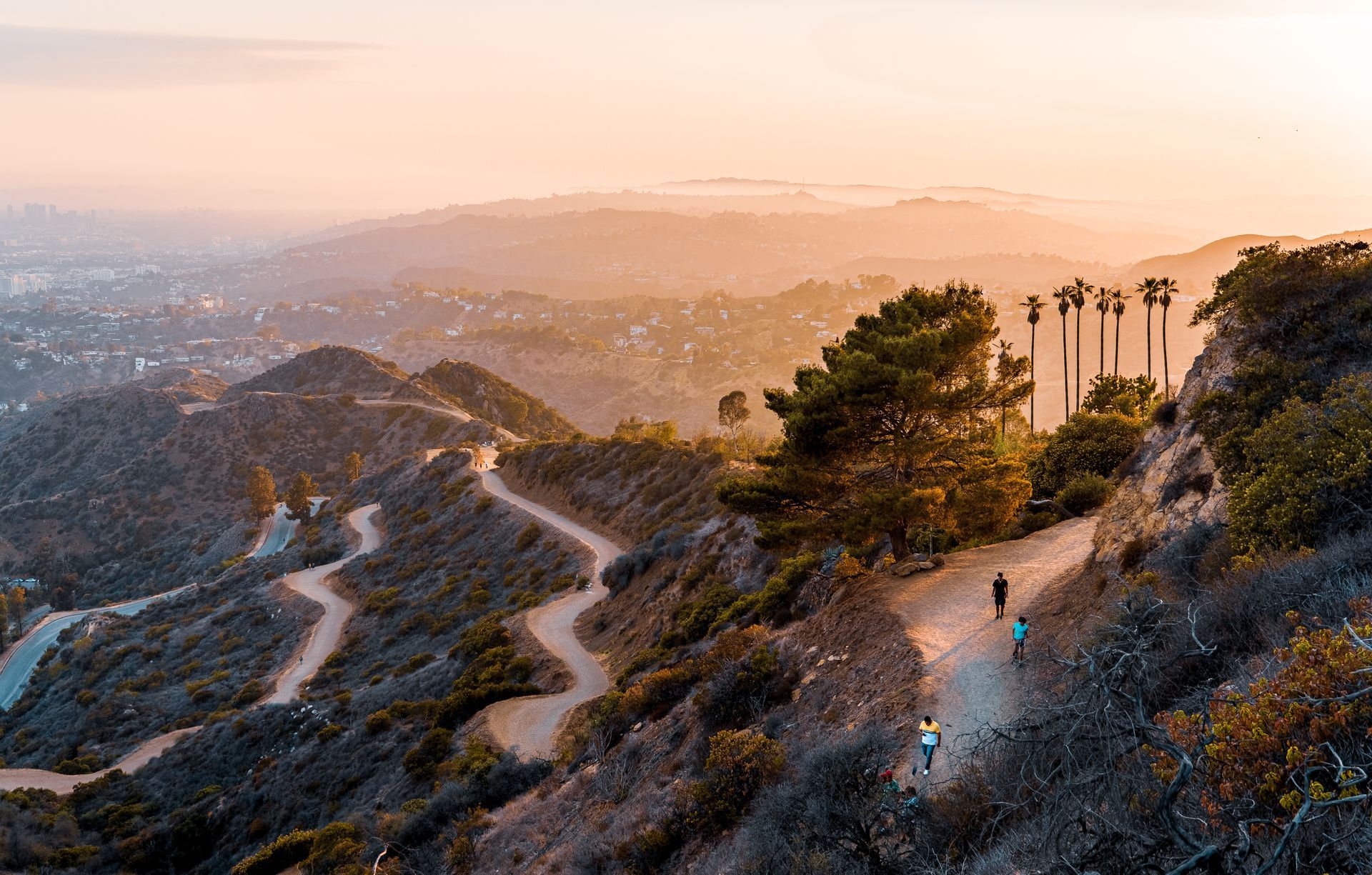Winding hiking trail on a hillside at sunset; two people hike on the path, with a distant city visible.