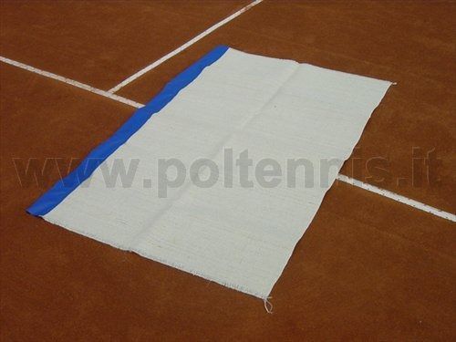 Un tappetino rettangolare di tessuto bianco con una striscia blu lungo un bordo, steso su un campo da tennis in terra battuta.