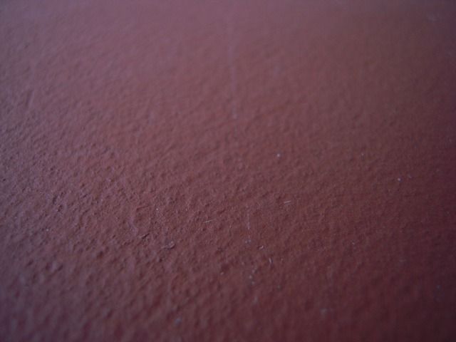 Primo piano di una superficie ruvida di una parete dipinta di color bordeaux scuro o rosso-brunastro.