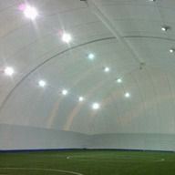 L'interno di una struttura sportiva coperta, con un campo in erba sintetica verde sotto un alto soffitto ad arco bianco e illuminato.