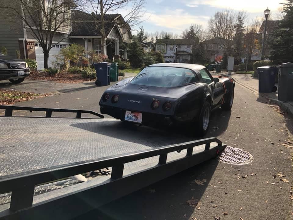 A black corvette is being towed down a street by a tow truck.