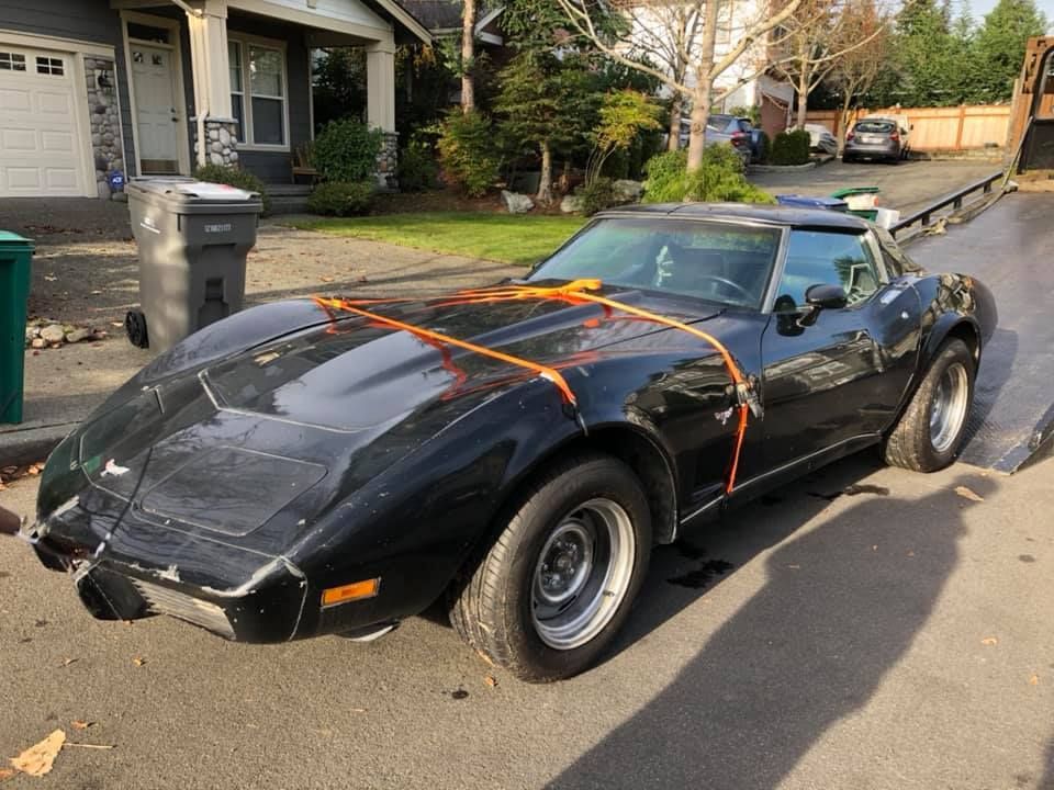 A black corvette is tied to a tow truck on the side of the road.