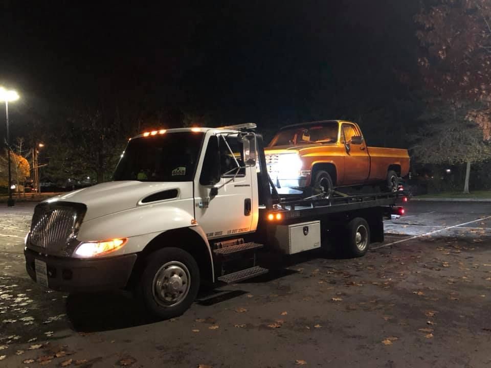 A tow truck is towing a yellow truck in a parking lot at night.