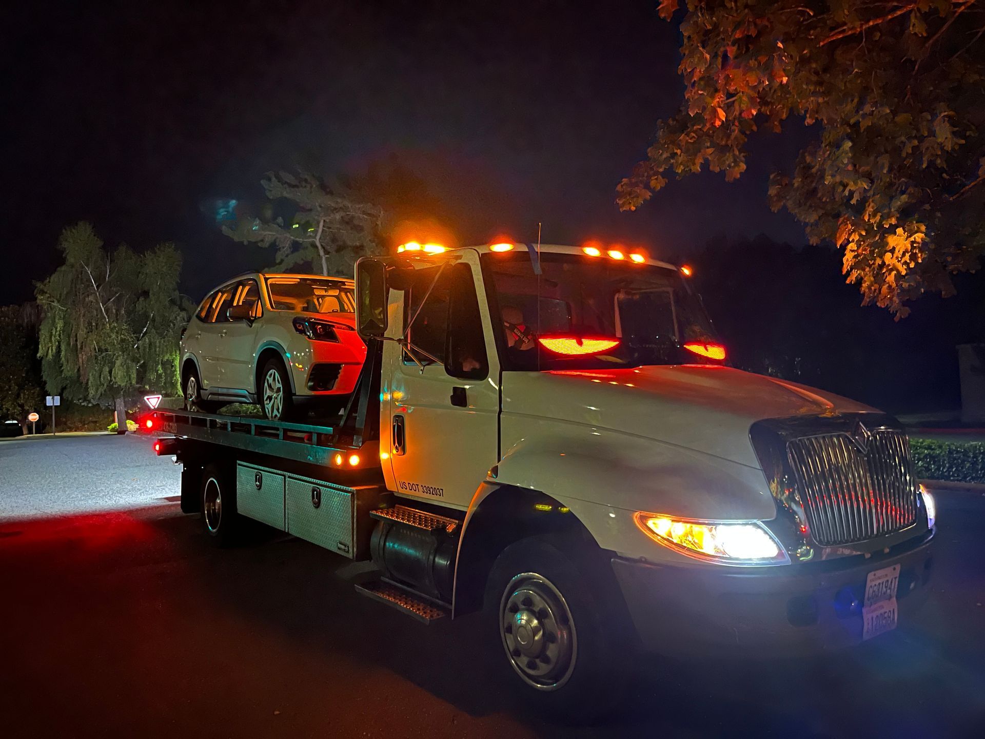 Tow truck at night hauling a white car; warning lights flashing.