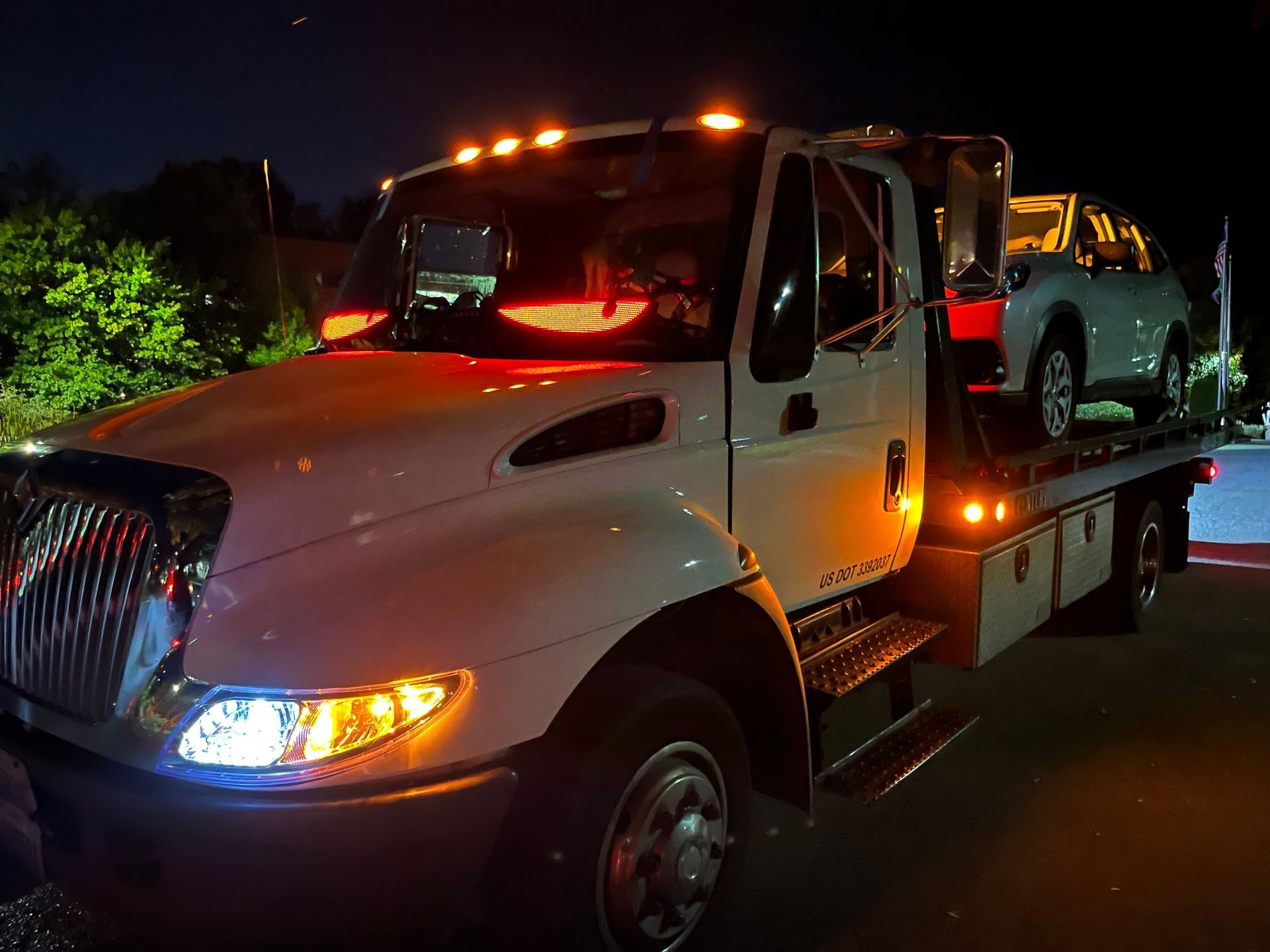 White tow truck with flashing lights at night, carrying a light-colored car.