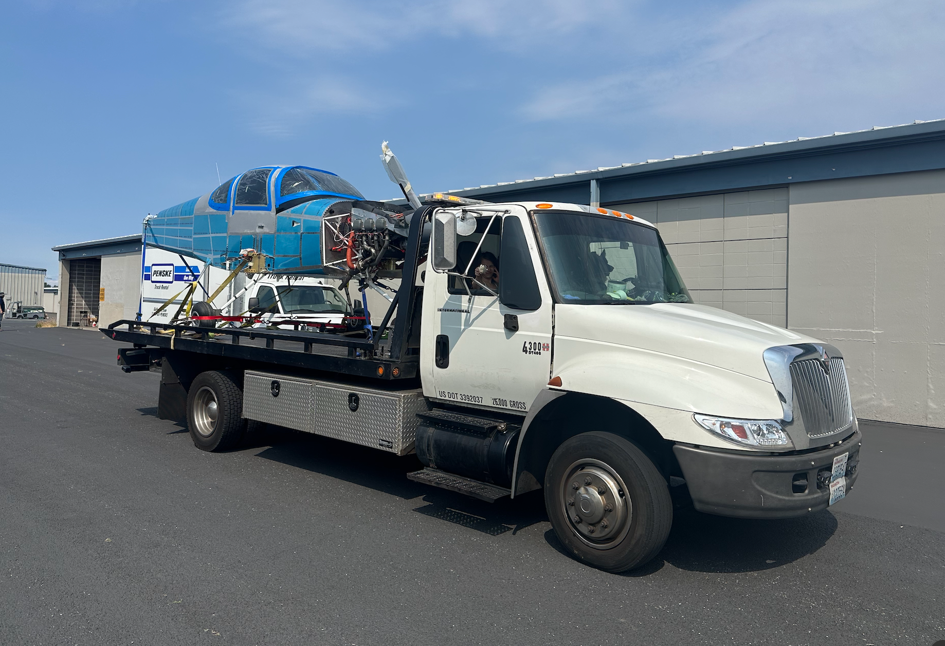 A white tow truck is carrying a blue airplane on the back.