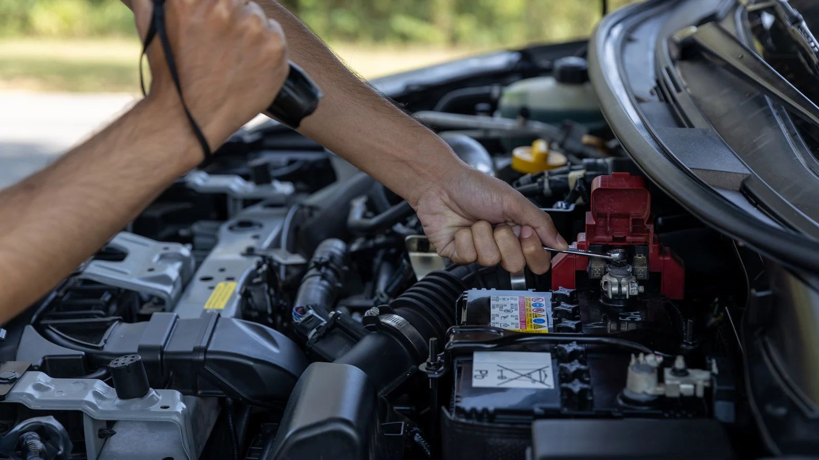 Person using a flashlight to inspect car engine, focusing on the battery and wiring.