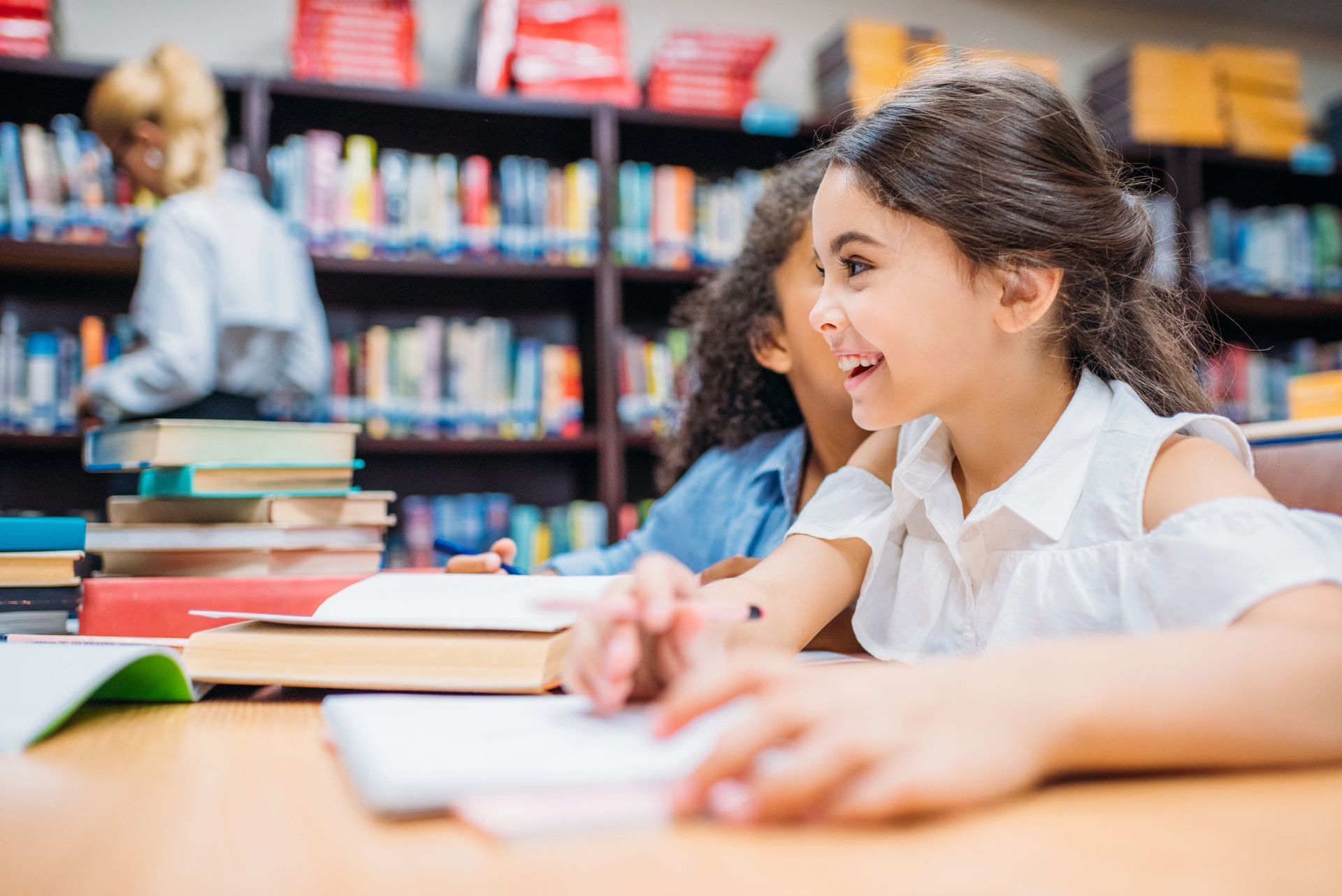 Dos estudiantes sonríen mientras estudian en una mesa de una biblioteca con estanterías al fondo.