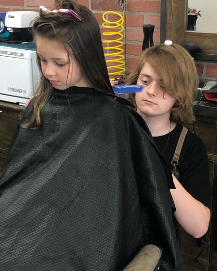 A stylist with brown hair cuts the long hair of a client wearing a black cape in a barbershop with a brick wall.
