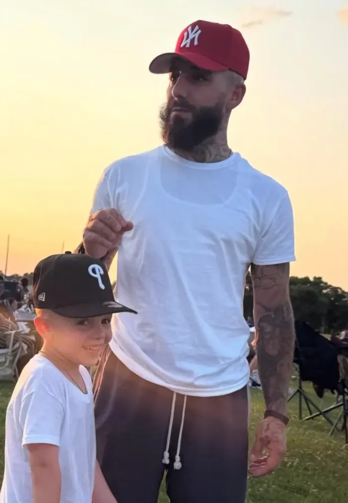 A bearded man in a red cap and a child in a black cap standing side-by-side in white shirts outdoors at sunset.