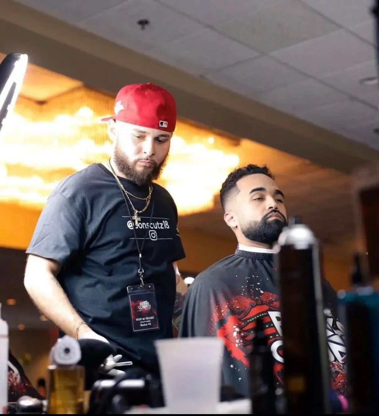 A barber wearing a red cap and black shirt focuses on grooming a client sitting in a salon chair.