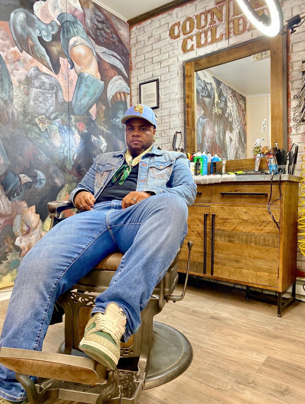 A person sits in a barbershop chair, wearing a light blue cap, denim jacket, jeans, and patterned sneakers.