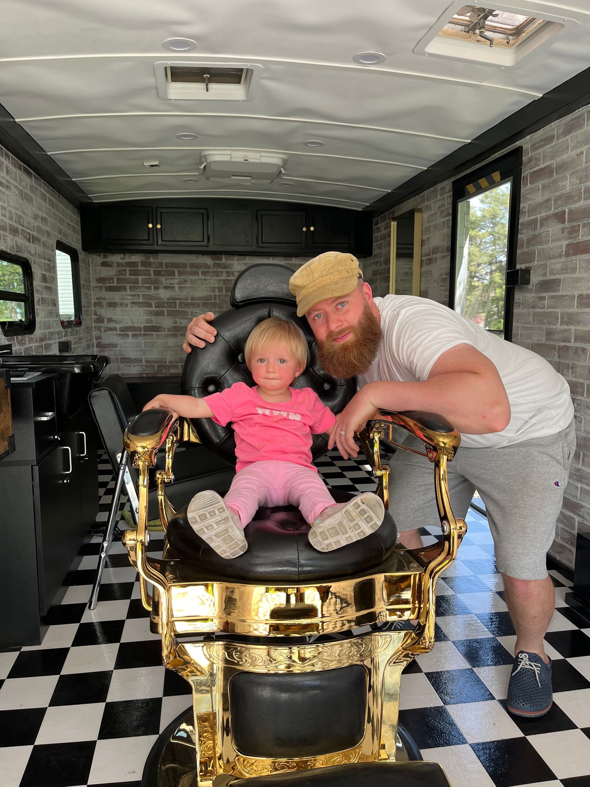 A child sits in a gold-accented barber chair inside a mobile shop with a black-and-white checkered floor and a bearded man.