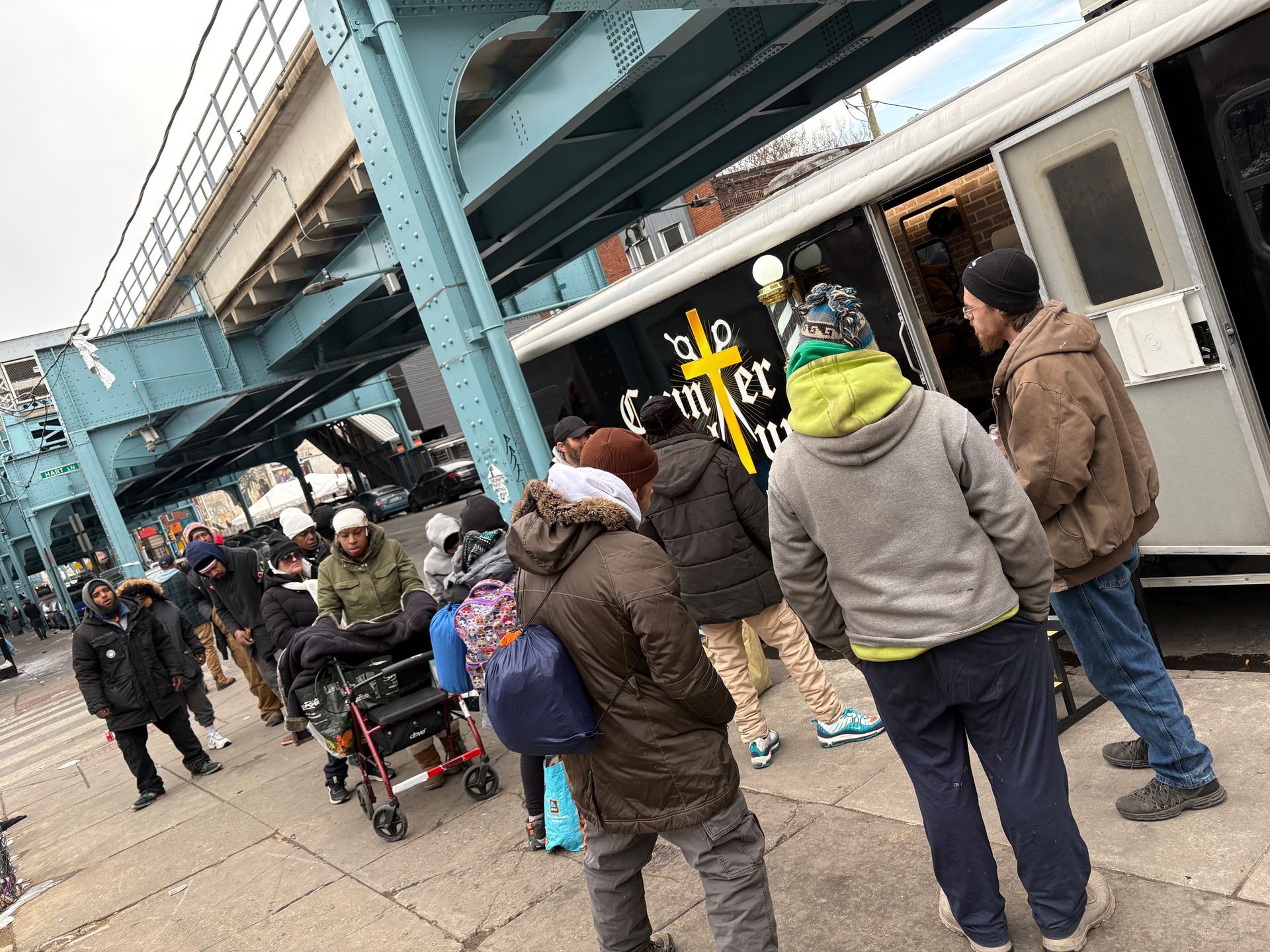 A queue of people in winter clothing stand outside a trailer displaying a yellow cross logo beneath an elevated train track.