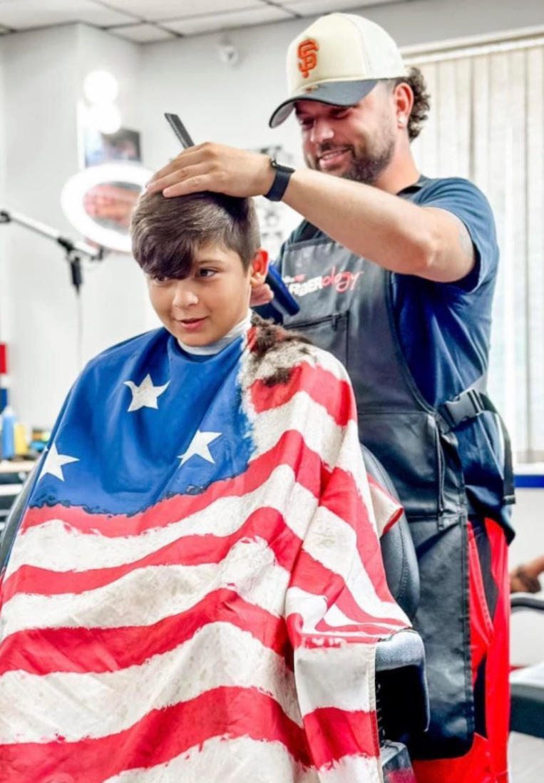 A barber with a baseball cap cuts a client's hair, while the client wears a cape with an American flag design.