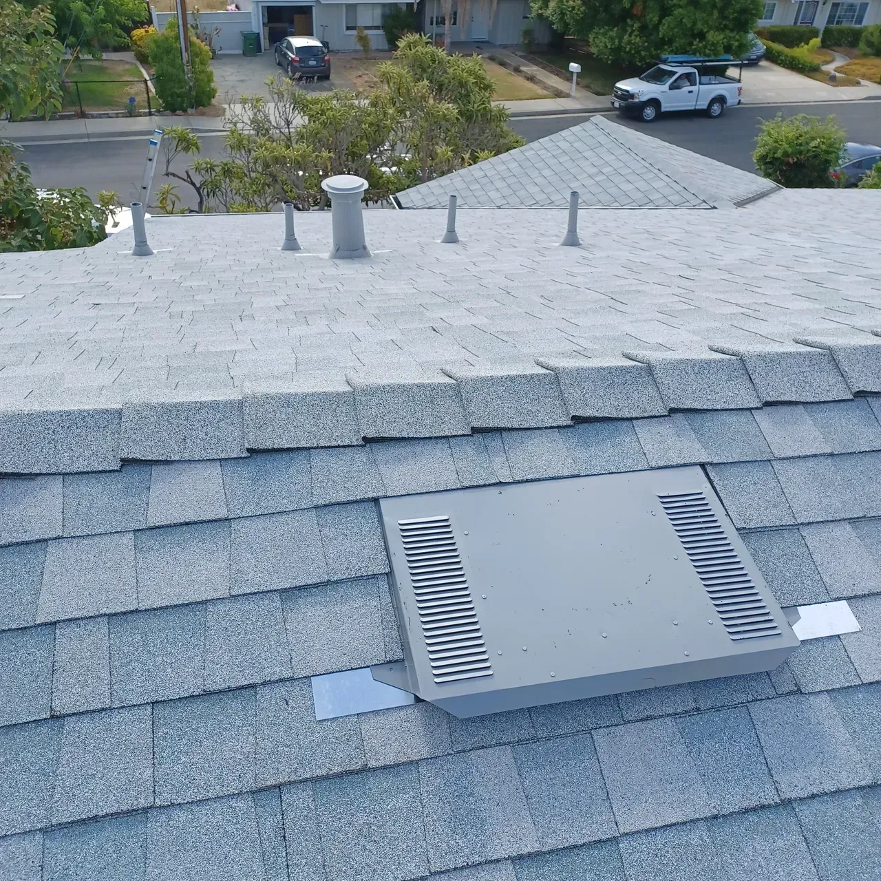 Gray shingle roof with a vent, several pipes, and a residential neighborhood in the background.