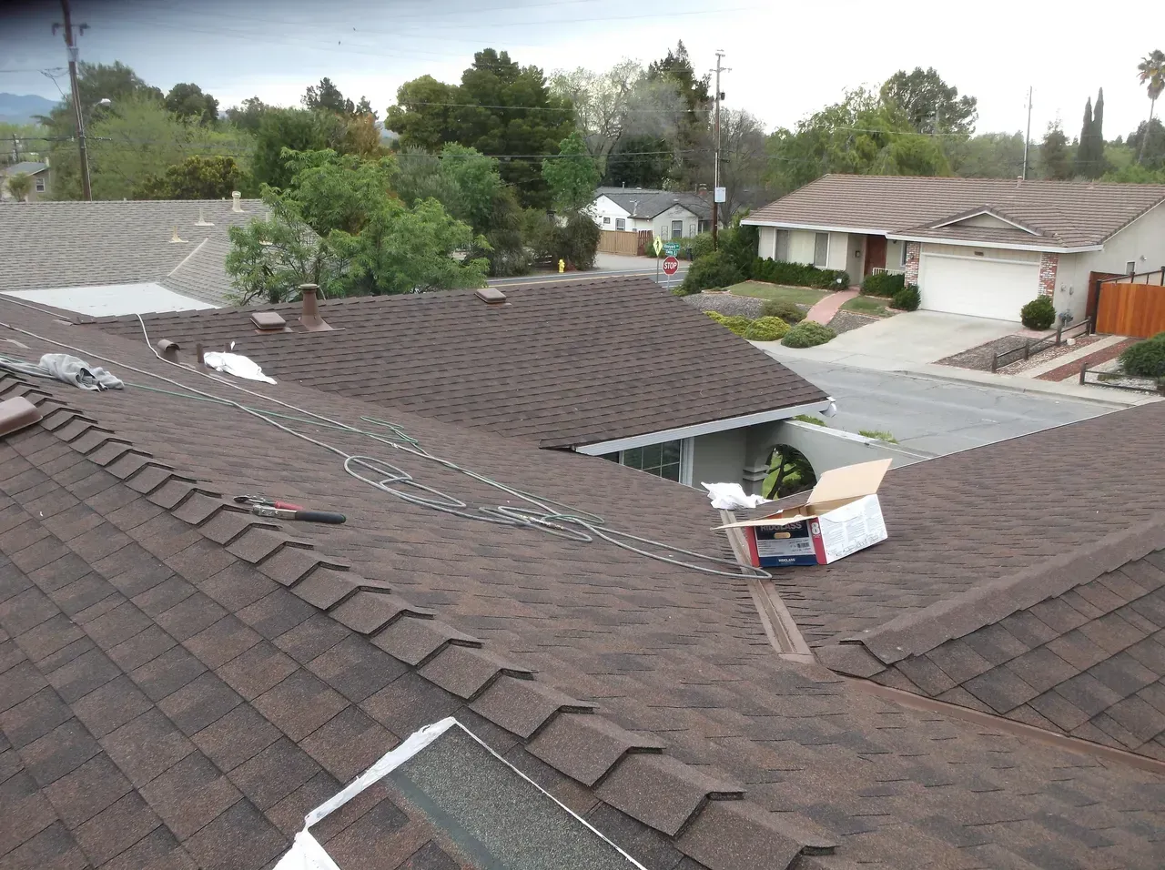 Brown shingled roofs in residential area with a view of houses, trees, and sky.