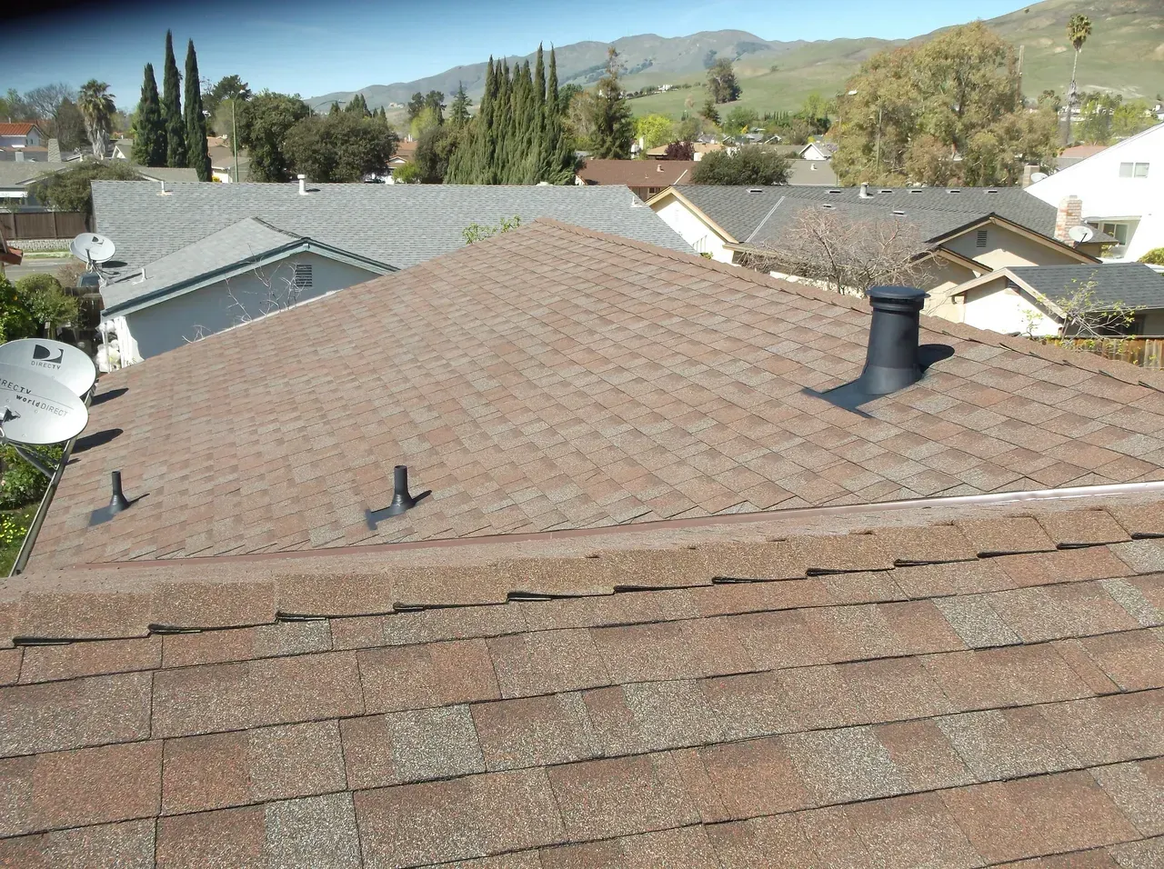 View of a brown shingle roof with a black vent. Houses and trees in the background.