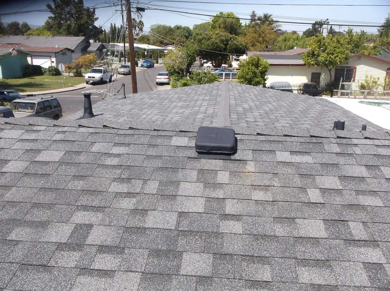 A gray shingle roof with a vent and a square device on top. Houses and trees in the background.