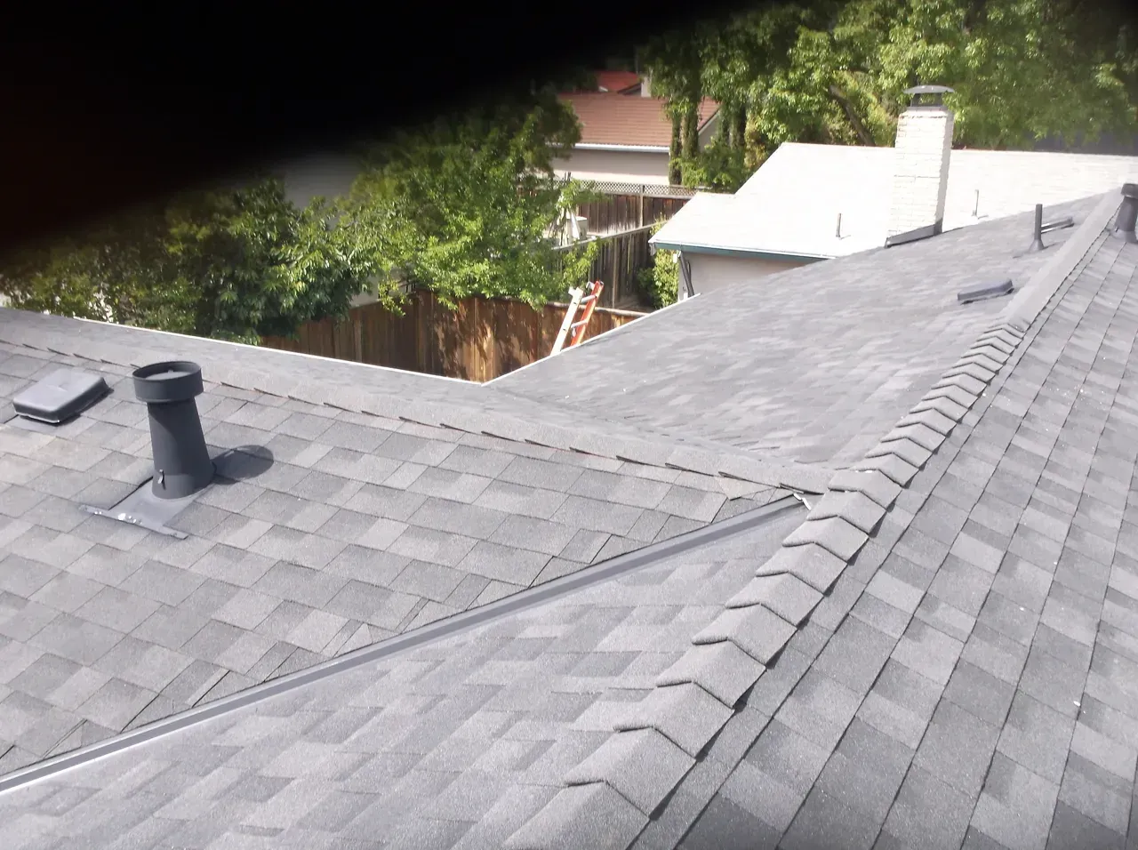 A gray shingle roof with a vent pipe, surrounding trees, and a house in the background.