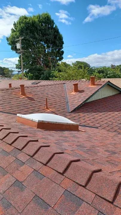 View of a roof with red and black asphalt shingles, vents, and a skylight under a blue sky.